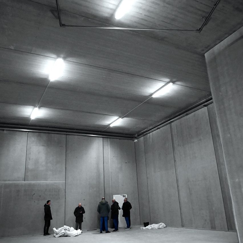 A monochromatic image of an empty room with concrete walls and ceiling. Four individuals are present, dressed in winter clothing. One person stands near a white object on the floor.
