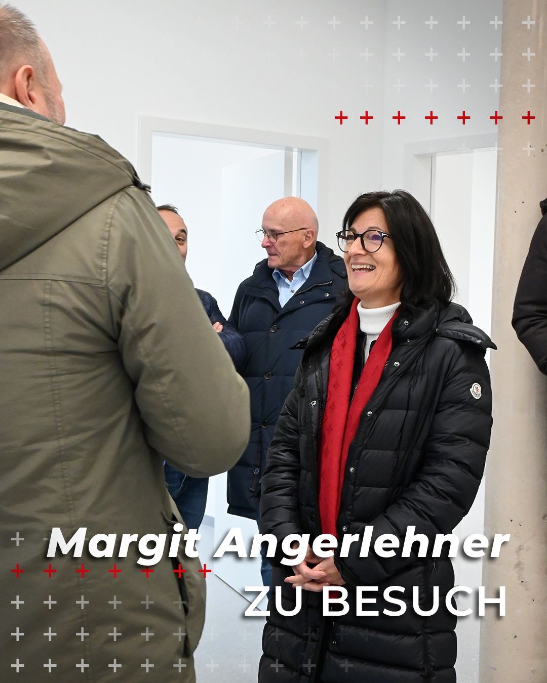 Margit Angerer, a woman wearing a black puffer jacket and a red scarf, stands smiling in a room with two men, one in a blue jacket and the other in a green jacket. The background is white with red plus signs.
