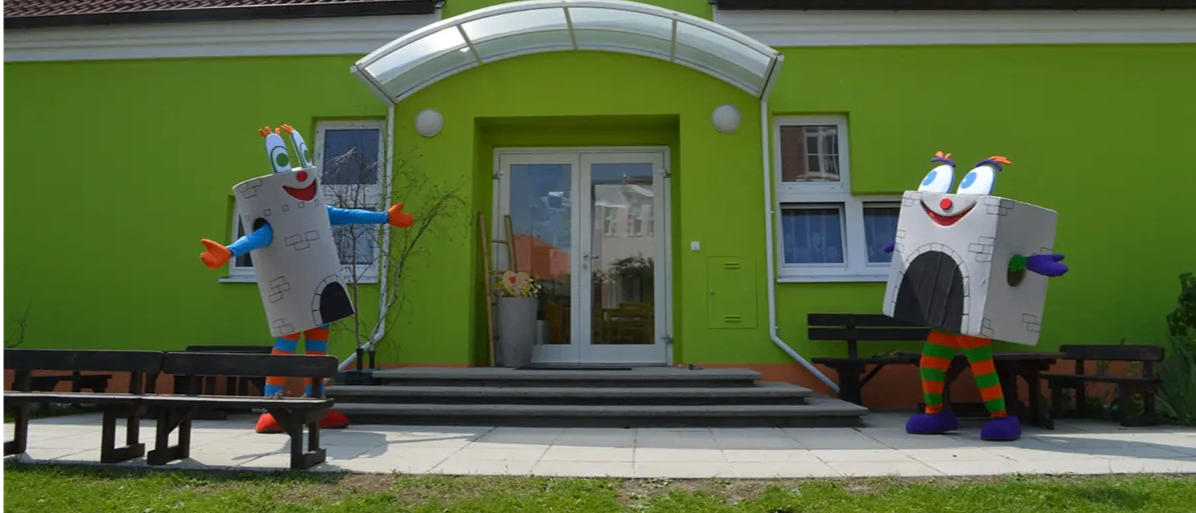 Zwei Maskottchen stehen vor einem grünen Gebäude mit einem braunen Dach. Eines trägt eine Turm-Kostüm, und das andere ein Burg-Kostüm. Der Eingang besteht aus doppelten Glastüren und einer kleinen Veranda.