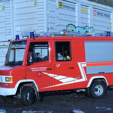 Ein roter Feuerwehrwagen mit blauen Lichtern auf dem Dach steht vor einem großen weißen Container mit Swiss Krono-Markierung.