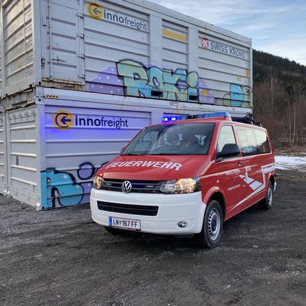 Ein roter Volkswagen-Transporter mit der Aufschrift 'Feuerwehr' steht vor einem großen Container mit Graffiti.