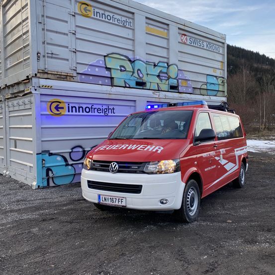 Ein roter Volkswagen-Transporter mit der Aufschrift 'Feuerwehr' steht vor einem großen Container mit Graffiti.