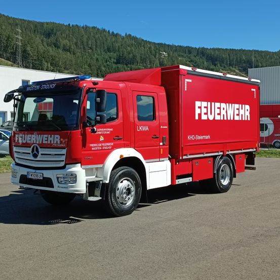Ein roter Mercedes-Benz Feuerwehrwagen mit 'FEUERWEHR' auf der Seite, in einem Parkplatz mit einem Gebirgshintergrund geparkt.
