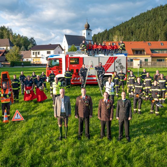 Eine Gruppe von Feuerwehrleuten steht vor einem Feuerwehrauto, auf dem einige sitzen, in Uniform gekleidet und posiert für ein Foto auf einem Feld mit einem Berg im Hintergrund.
