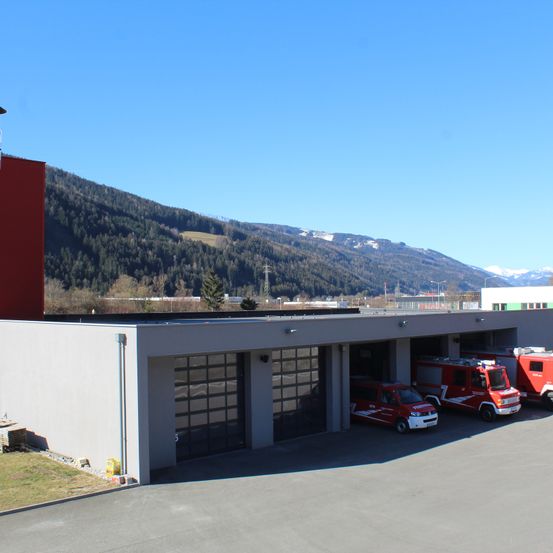 Eine Feuerwehrstation mit roten Fahrzeugen unter einer Garage, dahinter ein roter Turm und Berge im Hintergrund.