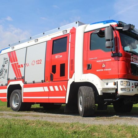 Ein roter Feuerwehrwagen mit der Aufschrift HLF 2500 und 122 ist auf einem Kiesweg geparkt. Der Wagen hat ein blaues und weißes Dach, einen roten Körper und weiße Streifen.