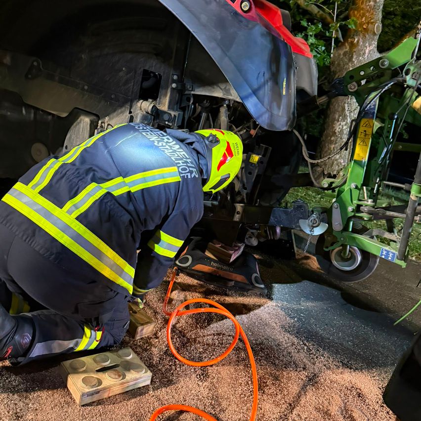 Ein Feuerwehrmann in voller Ausrüstung kniet neben einem großen Fahrzeug, möglicherweise einem Traktor, und arbeitet an dessen Unterboden. Die Szene spielt im Freien mit Kies und einem Baum in der Nähe.