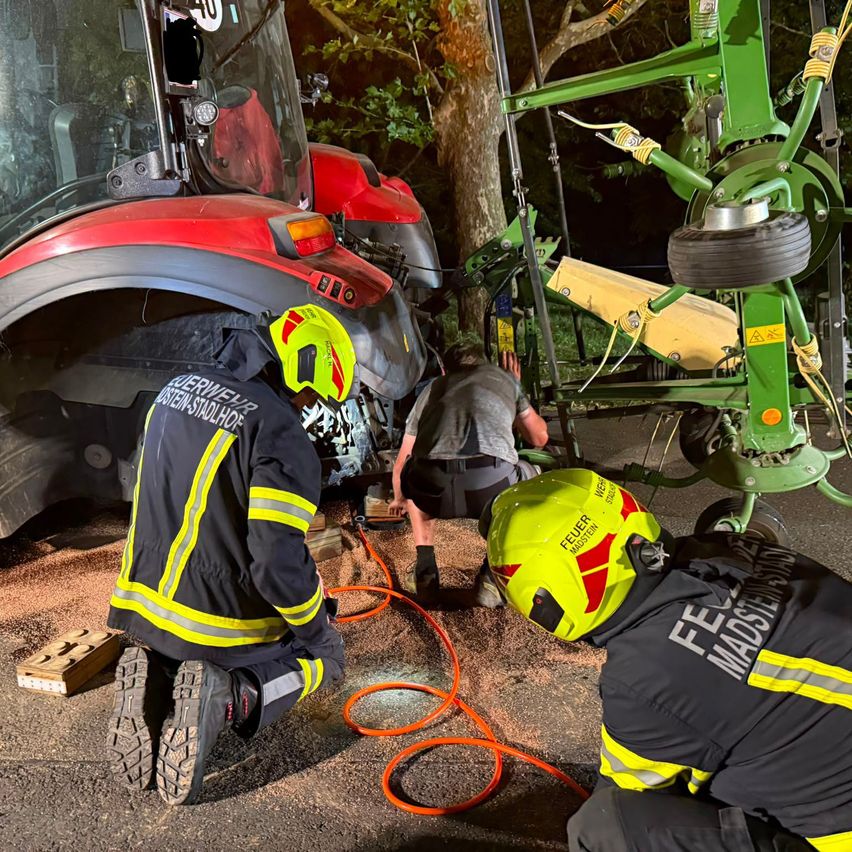 Feuerwehrleute in Schutzkleidung arbeiten an einem roten Traktor mit zerbrochener Windschutzscheibe und beschädigter Front. Ein grünes Maschinenfahrzeug ist in der Nähe.