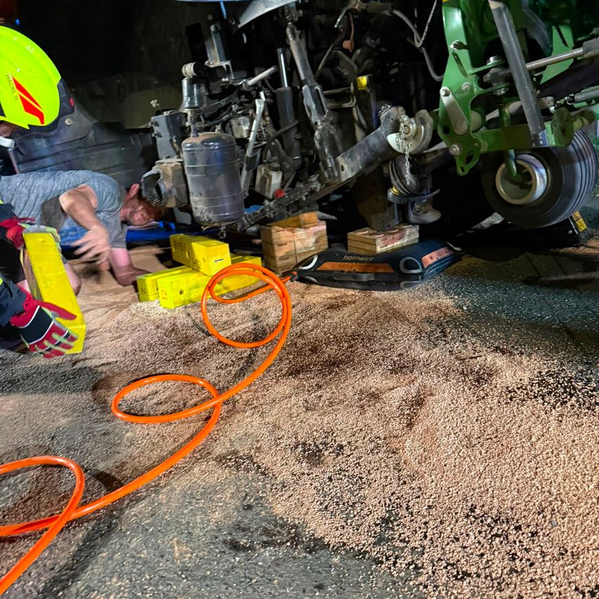 Zwei Arbeiter in Schutzkleidung arbeiten an einem großen Maschinenfahrzeug, wobei einer ein gelbes Kästchen hält und der andere sich beugt. Das Fahrzeug wird mit Hilfe eines hydraulischen Wagenhebers angehoben, und es gibt mehrere gelbe Kisten und einen orangefarbenen Schlauch auf dem Boden.