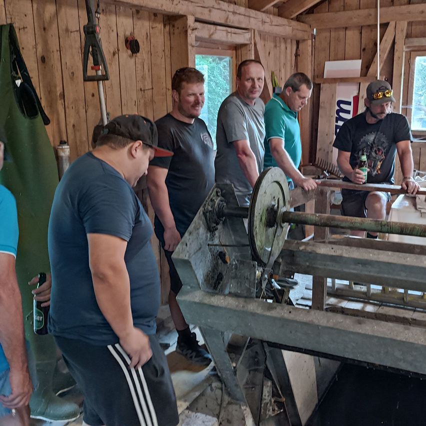 Fünf Männer in einer Werkstatt, einer hält ein Bier, ein anderer bedient eine Maschine. Holzwände und Werkzeuge im Hintergrund.