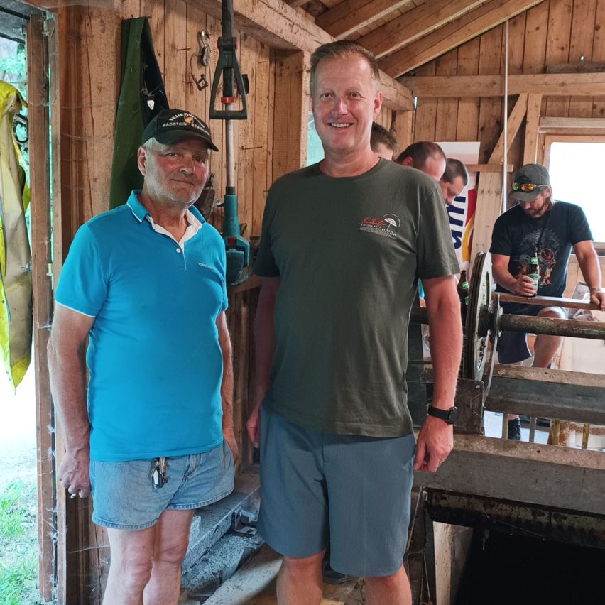 Zwei Männer stehen in einem Holzschuppen, einer trägt ein blaues Poloshirt und der andere ein grünes T-Shirt. Sie lächeln und scheinen für ein Foto zu posieren. Hinter ihnen hält ein Mann eine Flasche und eine weitere Person steht in der Nähe eines Fensters.