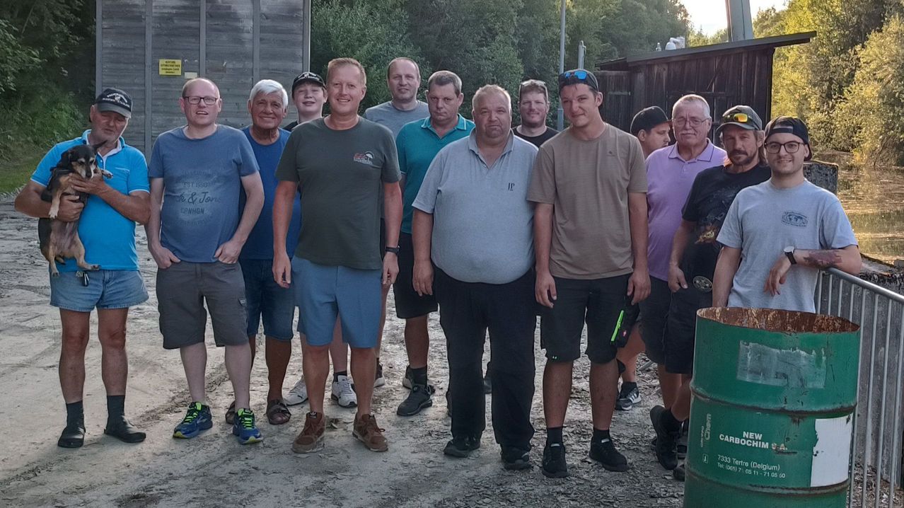 Eine Gruppe von Männern steht vor einem Holzgebäude, einige tragen Hüte und Brillen, mit einem grünen Fass im Hintergrund.