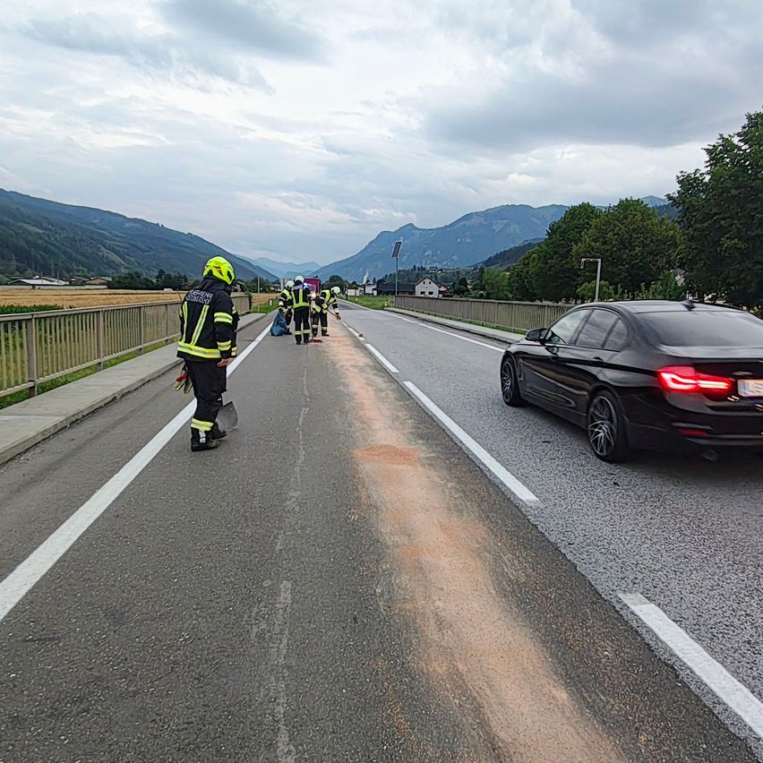 Eine Gruppe von Feuerwehrleuten reinigt eine Straße, an der ein schwarzes Auto vorbeifährt. Die Straße hat weiße Markierungen und eine Metallschiene an der Seite.