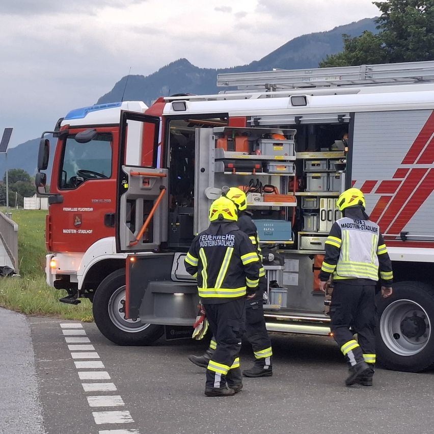 Drei Feuerwehrleute in gelben und schwarzen Uniformen verlassen einen Feuerwehrwagen mit offener Tür. Der Wagen ist am Straßenrand geparkt, mit einem Grasbereich und Bergen im Hintergrund.