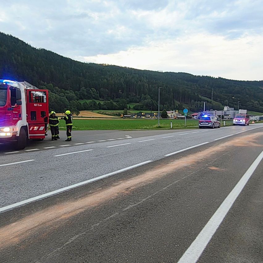 Zwei Feuerwehrleute stehen neben einem roten Feuerwehrauto am Straßenrand mit einem Berg im Hintergrund.