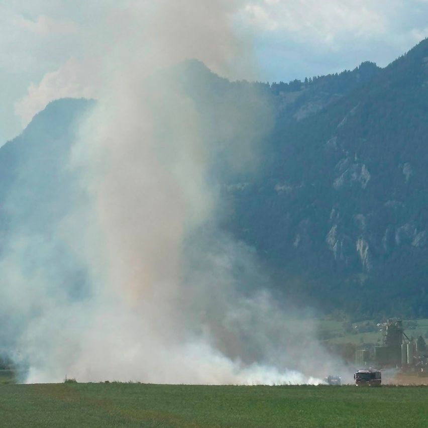 A large plume of smoke rises from a field, with mountains and a building in the background.