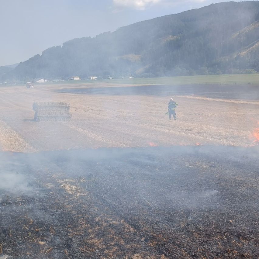 A firefighter stands in a field with a burning haystack and smoke rising, with a mountain and village in the background.