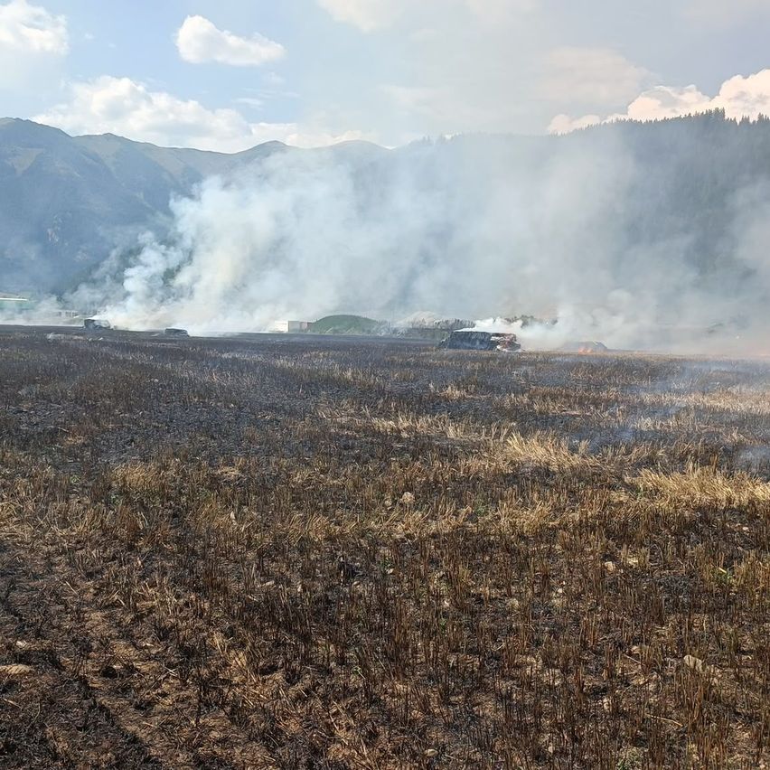 A wide, burnt field with smoke rising from the ground and a distant mountain range under a partly cloudy sky.