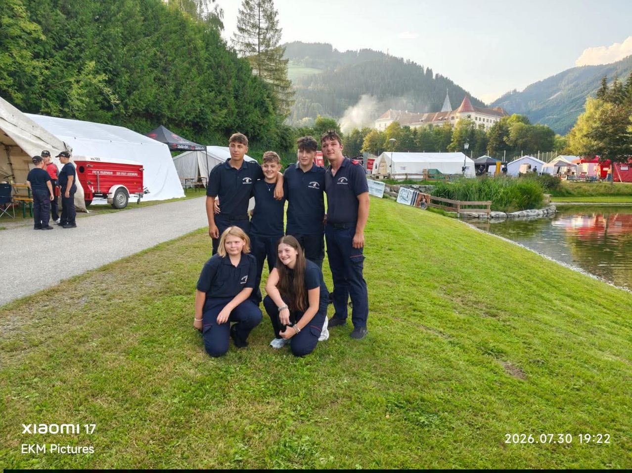 Five individuals in blue uniforms pose for a photo on a grassy field with mountains and a lake in the background.