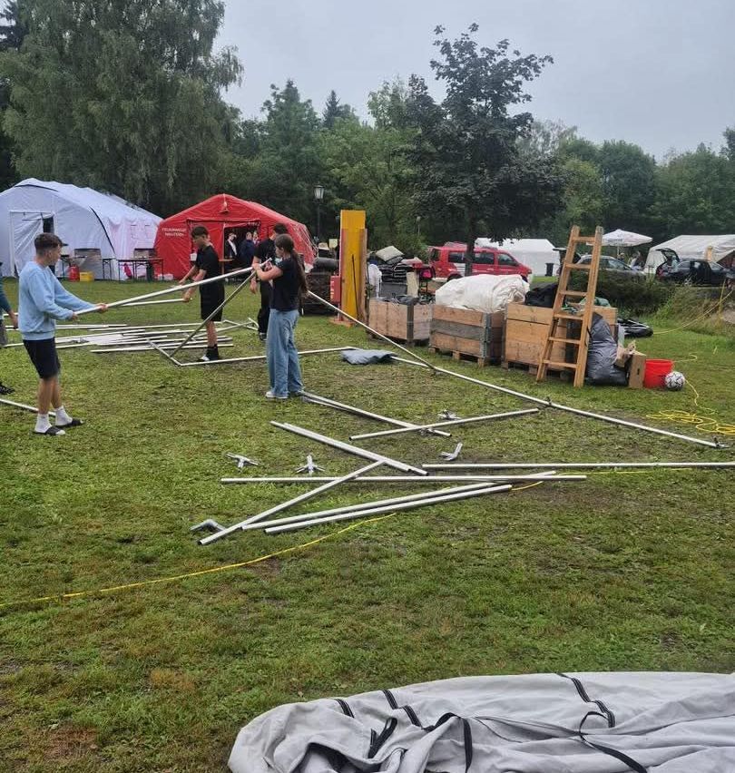 A group of people are setting up a tent on a grassy field with various tents and vehicles in the background.