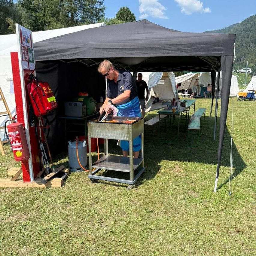 A man is grilling food under a black tent with a fire extinguisher and other equipment nearby.