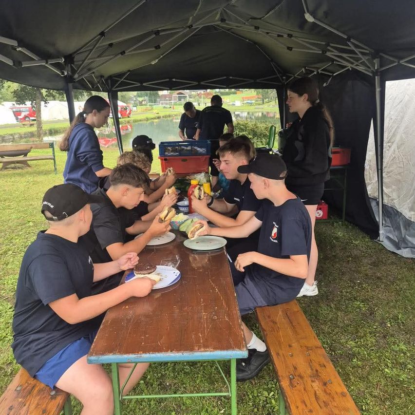 A group of young people are sitting around a table under a tent, eating sandwiches. They are wearing matching uniforms.