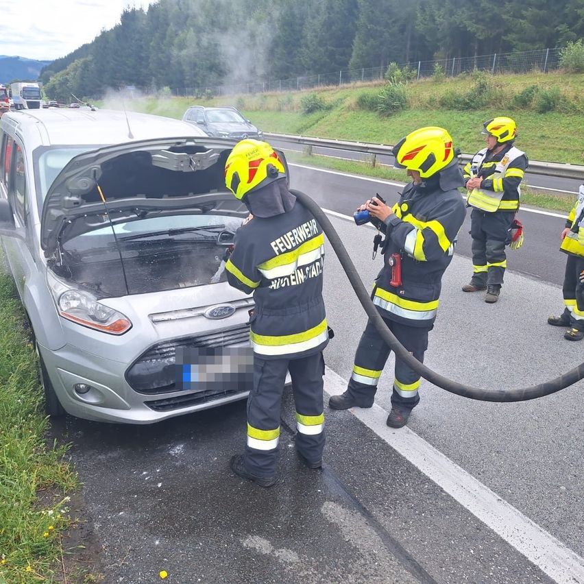Feuerwehrleute arbeiten an einem silbernen Ford-Fahrzeug am Straßenrand. Ein Feuerwehrmann benutzt einen Schlauch, um Wasser in den Motorraum des Autos zu sprühen.