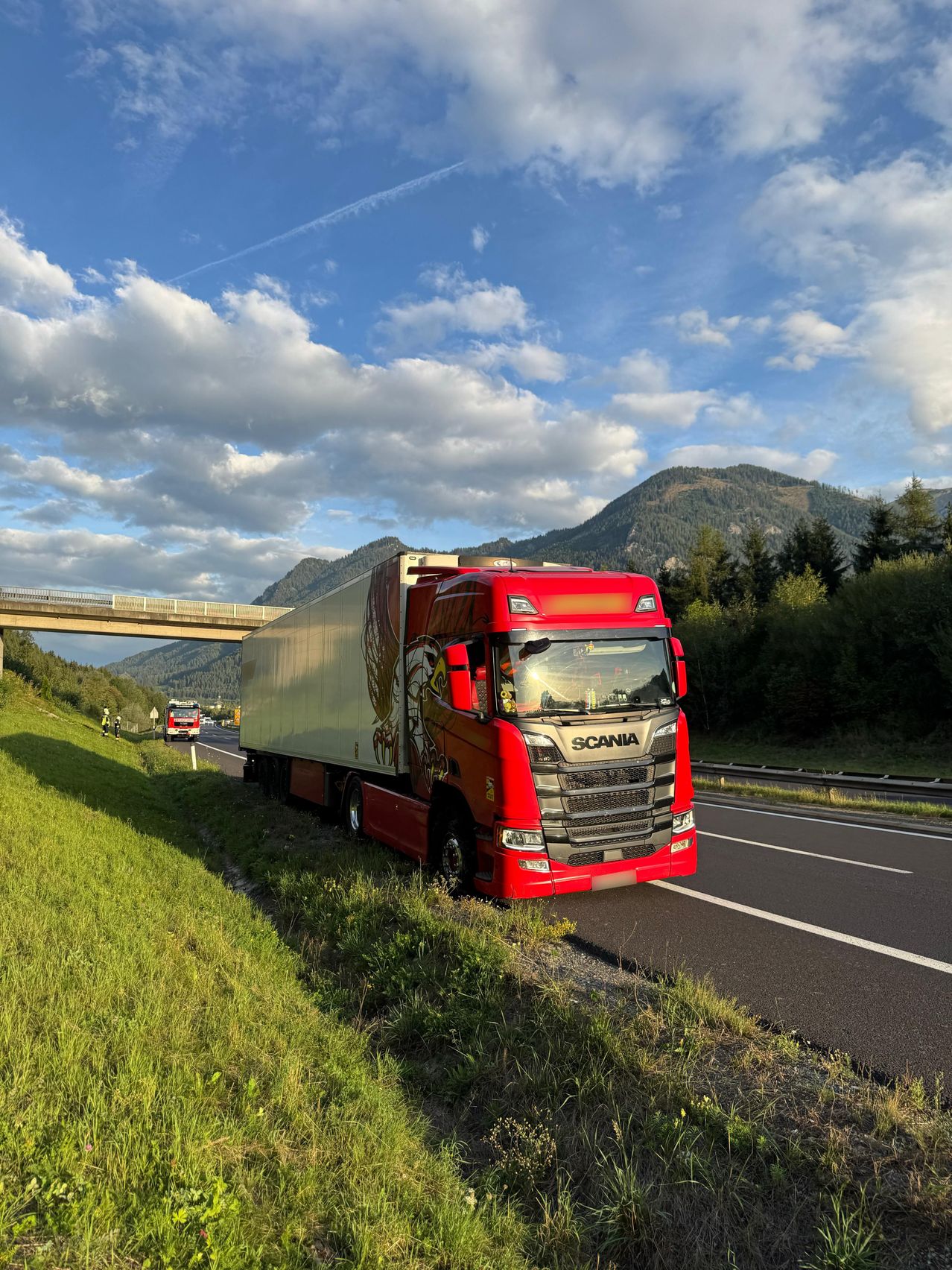 Ein roter Scania-Lkw ist am Straßenrand geparkt, mit einer Brücke und Bergen im Hintergrund.
