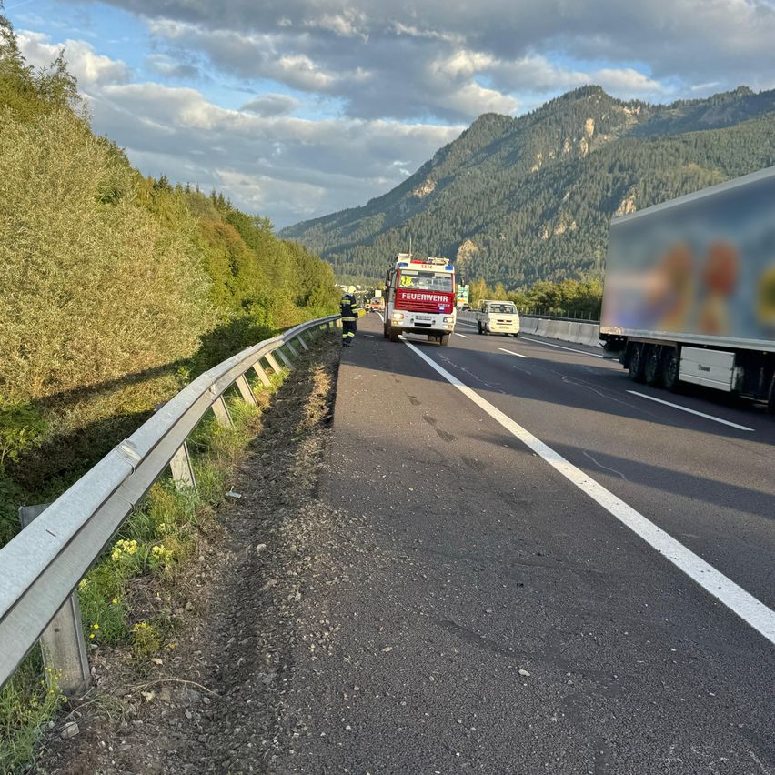 Eine Autobahn mit einem Feuerwehrauto und einer Person, die in der Nähe der Leitplanke steht. Ein Van und ein LKW fahren auf der Straße. Im Hintergrund sind Berge zu sehen.