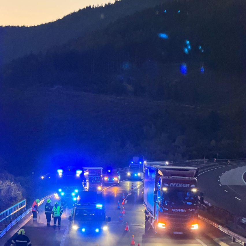 Notfallfahrzeuge mit blauen Blinklichtern sind auf einer Autobahn in der Nacht geparkt. Arbeiter in reflektierenden Westen stehen in der Nähe der Fahrzeuge. Die Straße ist von Bergen und Bäumen umgeben.