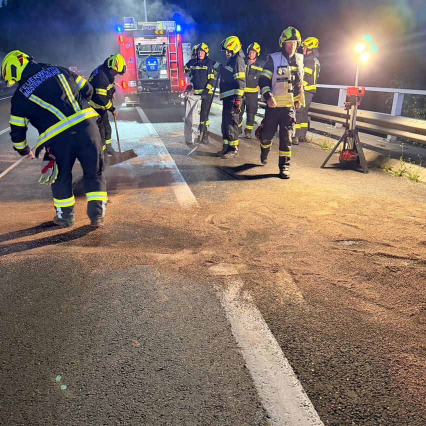 Feuerwehrleute reinigen eine Straße bei Nacht, mit einem Feuerwehrauto und Notfallleuchten im Hintergrund.