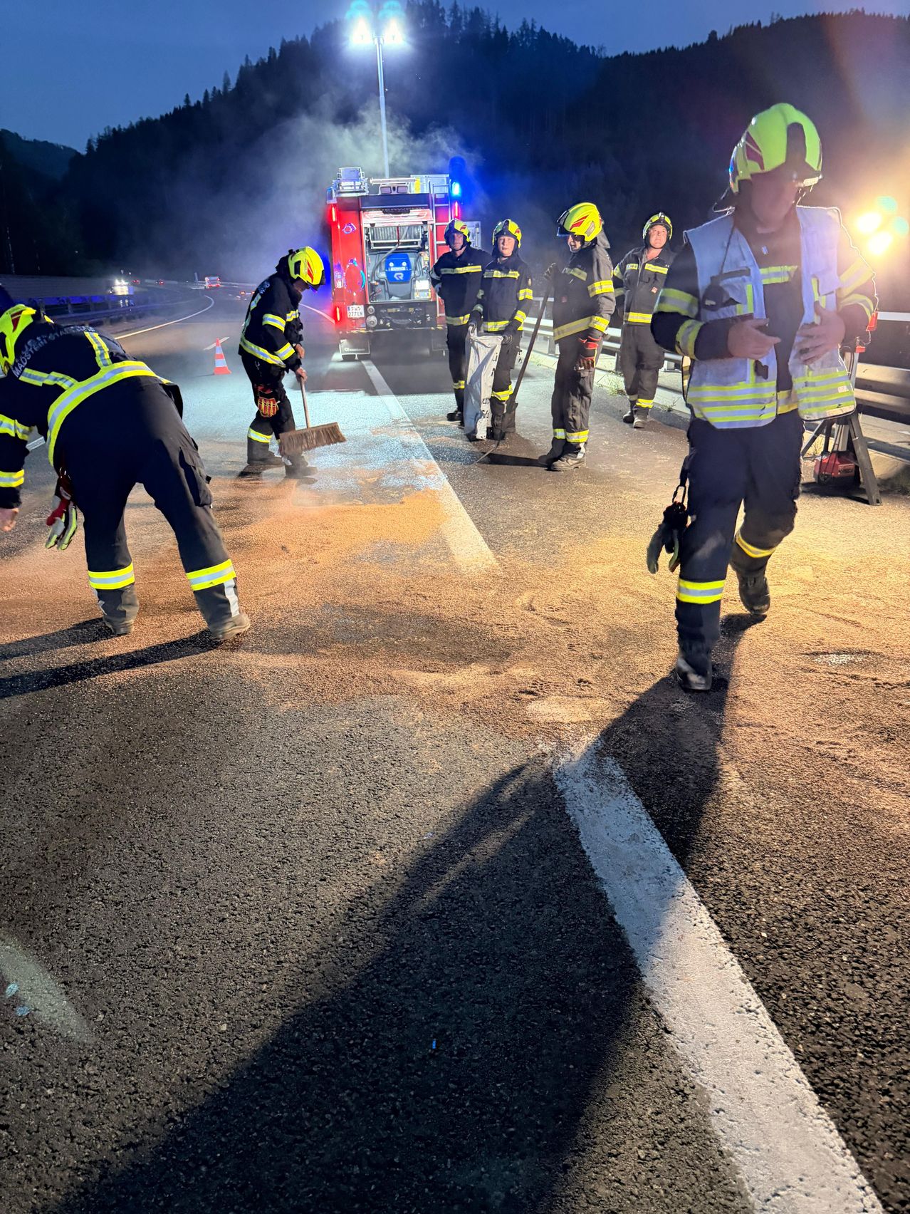Feuerwehrleute reinigen die Straße bei Nacht, beleuchtet von blinkenden Lichtern und Rauch.