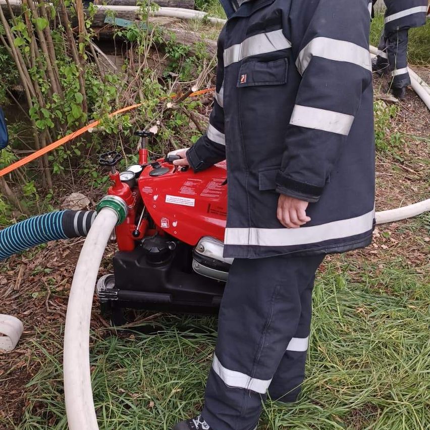 Ein Feuerwehrmann steht neben einer roten Maschine und hält einen Schlauch, der daran angeschlossen ist. Ein anderer Feuerwehrmann ist teilweise im Hintergrund zu sehen.