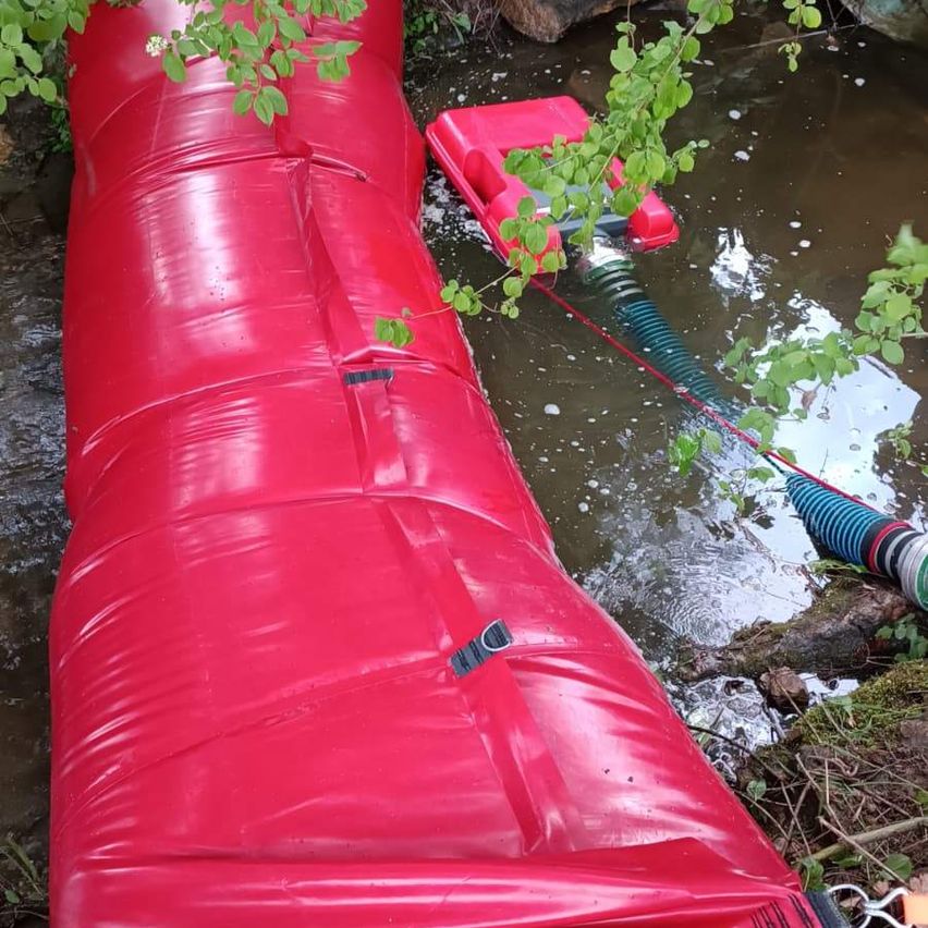Ein roter aufblasbarer Schlauch liegt in einem Gewässer. Ein rotes Objekt ist angeschlossen, verbunden mit einem Schlauch. Das Gebiet ist von Grün umgeben.