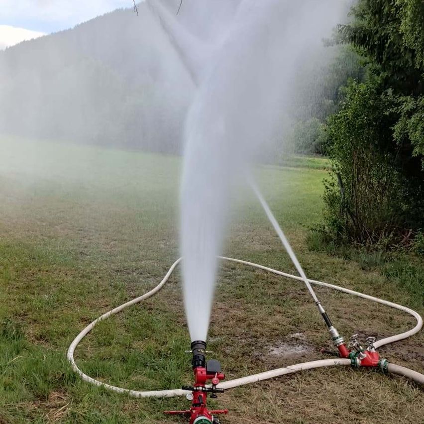 Zwei rote Feuerwehrschläuche spritzen Wasser in einem Außenbereich. Die Schläuche sind an eine Wasserquelle angeschlossen. Bäume und ein Berg sind im Hintergrund.