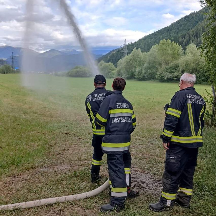 Drei Feuerwehrleute stehen auf einem Feld. Einer sprüht Wasser aus einem Schlauch, während die anderen zusehen. Der Himmel ist teilweise bewölkt, mit Bergen im Hintergrund.