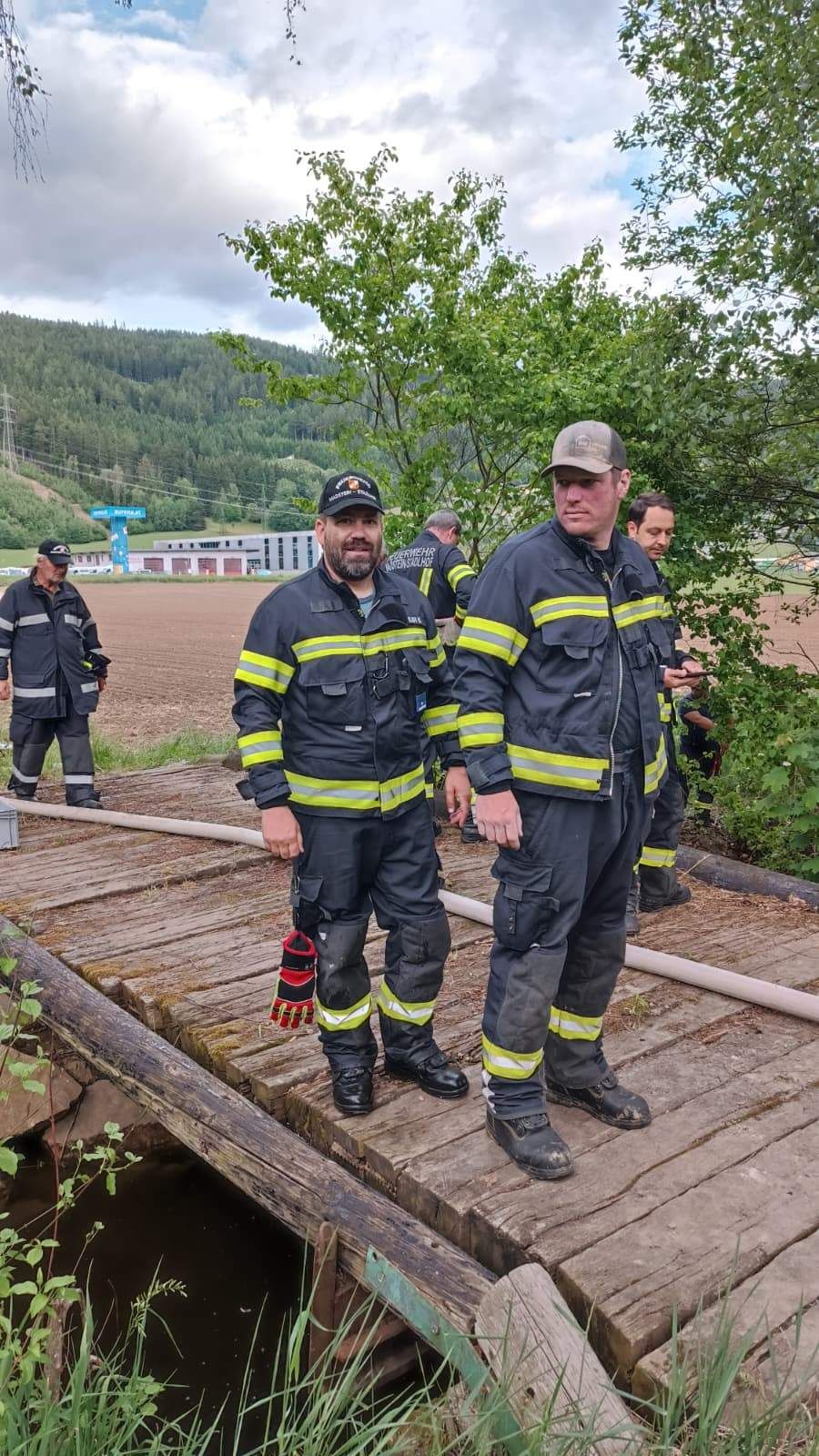 Feuerwehrleute in Uniform stehen auf einer Holzplattform mit Bäumen und Bergen im Hintergrund.