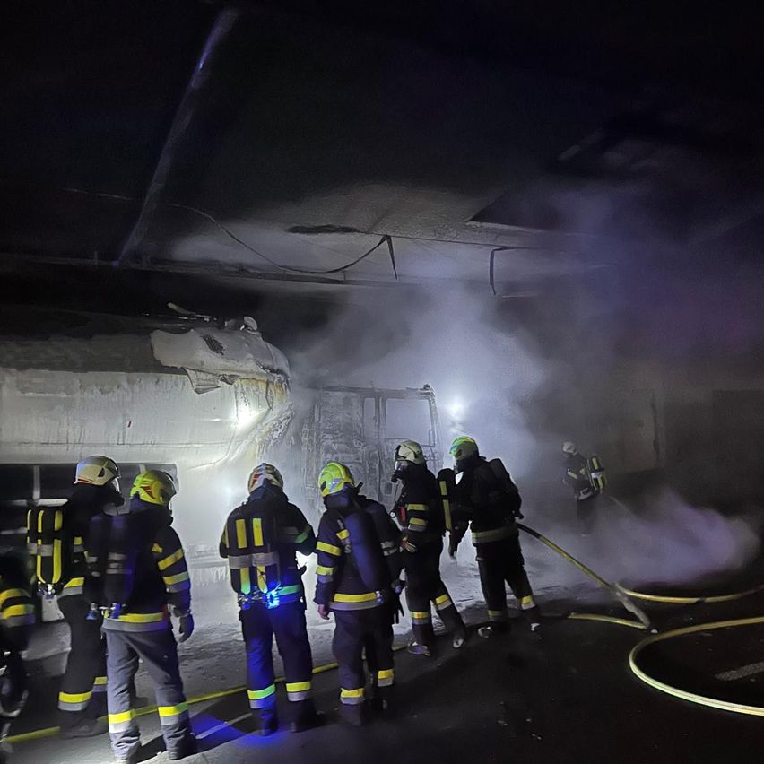 Feuerwehrleute in Schutzkleidung sind bei einem brennenden Gebäude in der Nacht und verwenden einen Schlauch, um Wasser zu sprühen. Rauch und Licht sind sichtbar.
