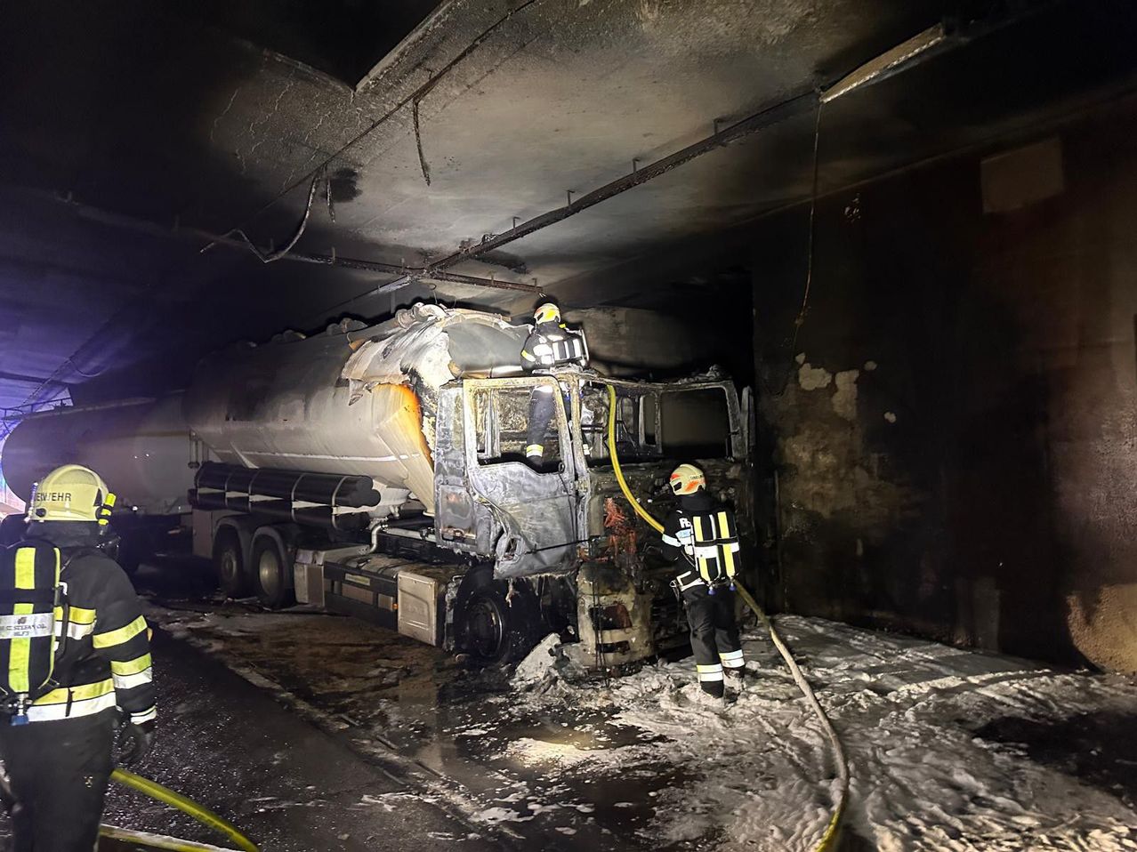 Feuerwehrleute löschen einen brennenden Tanklastwagen in einem Tunnel, einer oben und einer spritzt Wasser auf das Fahrzeug.