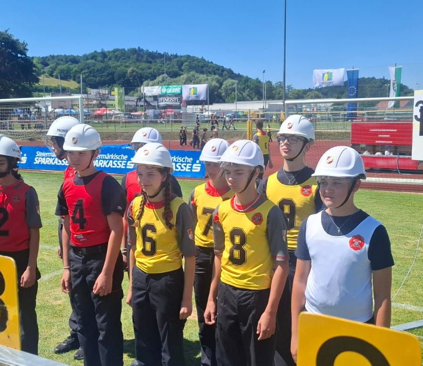 A group of young people in yellow and white uniforms with numbers are standing in a line on a field, wearing helmets and glasses. Behind them, a blurred background shows a fence, a flag, and trees.