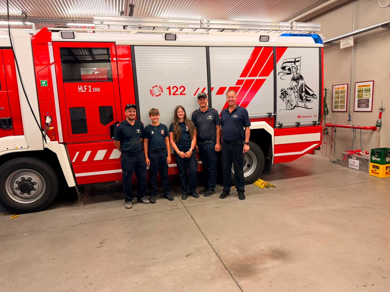 Five firefighters stand in front of a red truck. The truck is labeled HLF 2 2500 and has the number 122 on the side.