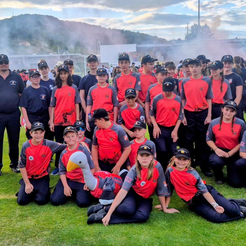 A team of young players and coaches in red and black uniforms poses for a photo on a grassy field with a mountain and buildings in the background.