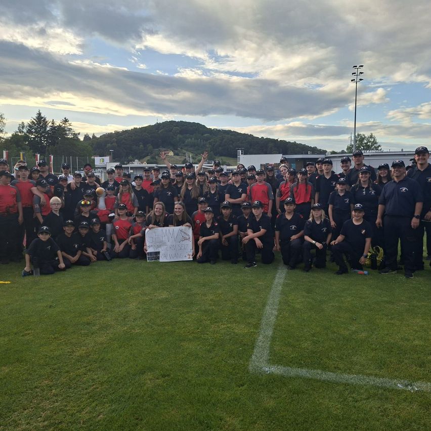 A group of people in matching uniforms poses for a photo on a sports field, with a mountain in the background and a sign held by some individuals.