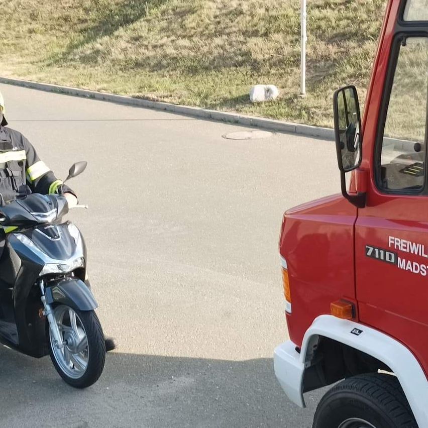 A firefighter on a scooter rides along a road, with a red truck visible in the background. The ground is paved, and there is some dry grass nearby.