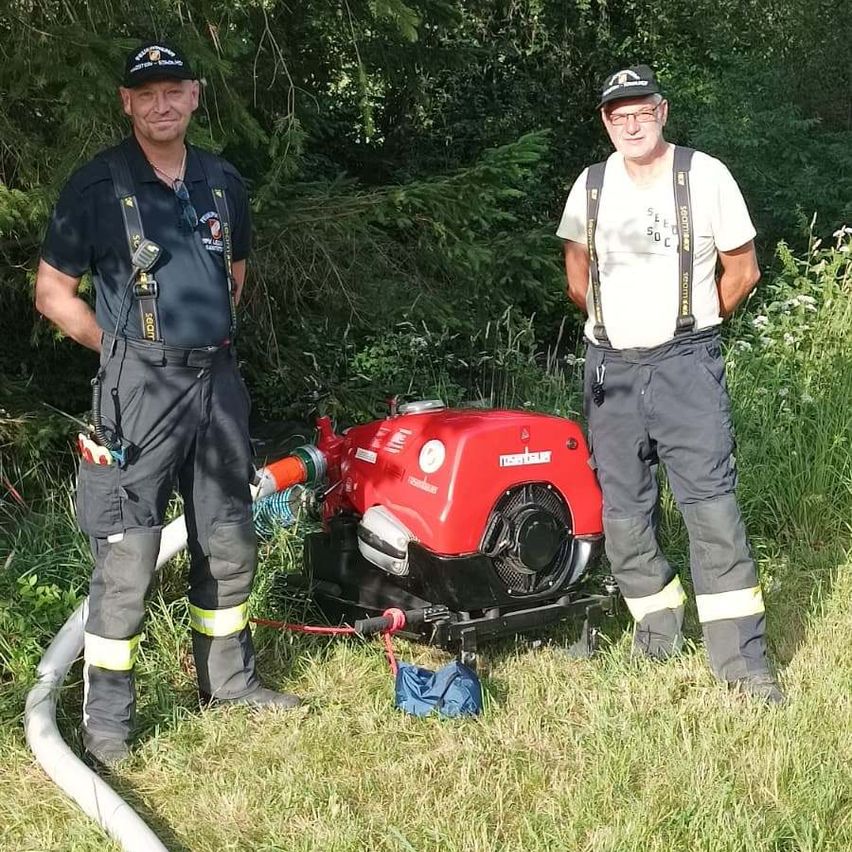 Two firefighters are standing outside with a red pump and hose, one wearing a hat and glasses and the other wearing a white shirt.