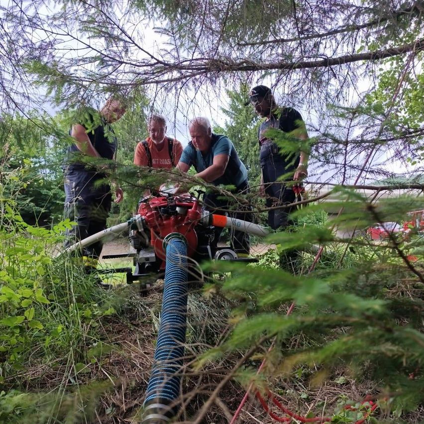 Four men in work gear work on a red machine with a long hose in a forest. Trees surround them.