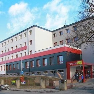 Ein modernes Gebäude mit mehreren Stockwerken, das mehrere Fenster und eine rot-graue Fassade aufweist. Menschen betreten das Gebäude durch den Eingang.