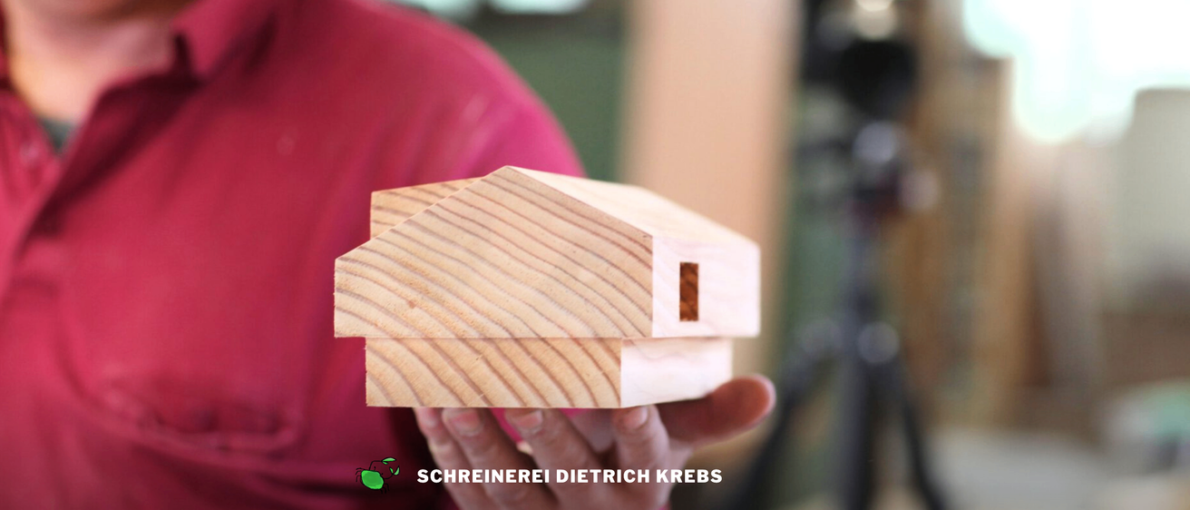 Eine Person hält ein hölzerne Modell eines Hauses mit dem Text 'SCHREINEREI DIETRICH KREBS' unten.