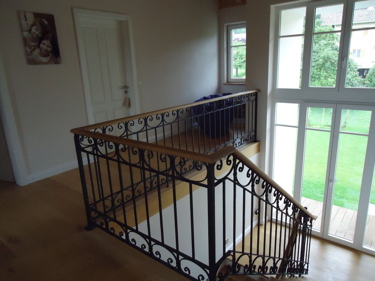 Eine moderne Treppe mit einem schwarzen Schmiedeeisengeländer und einem hölzernen Handlauf. Die Treppe führt zu einem Balkon mit Blick auf einen Rasen.
