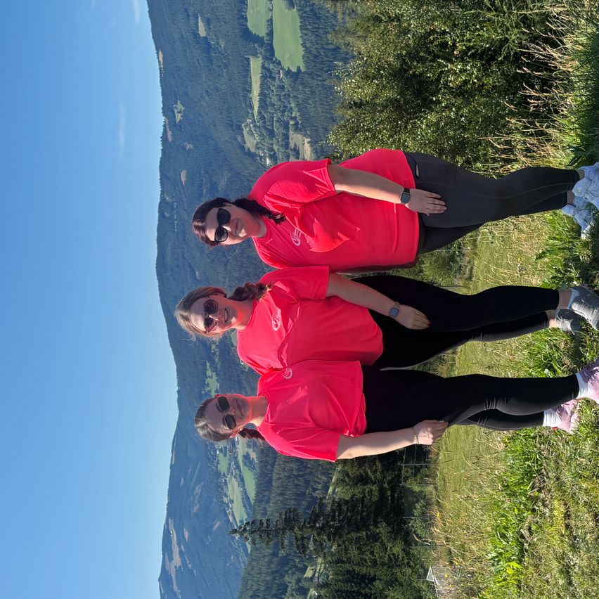 Drei Frauen in roten Shirts und schwarzen Leggings posieren für ein Foto auf einem grasbewachsenen Hügel mit Bergen im Hintergrund.